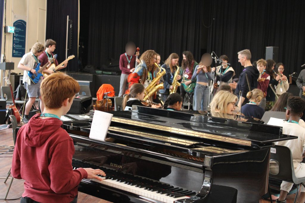 A group of young people in a music rehearsal space buzzes with energy as an orchestra of musicians comes together, featuring talents on piano, saxophones, and other instruments. A black grand piano is prominently in the foreground.